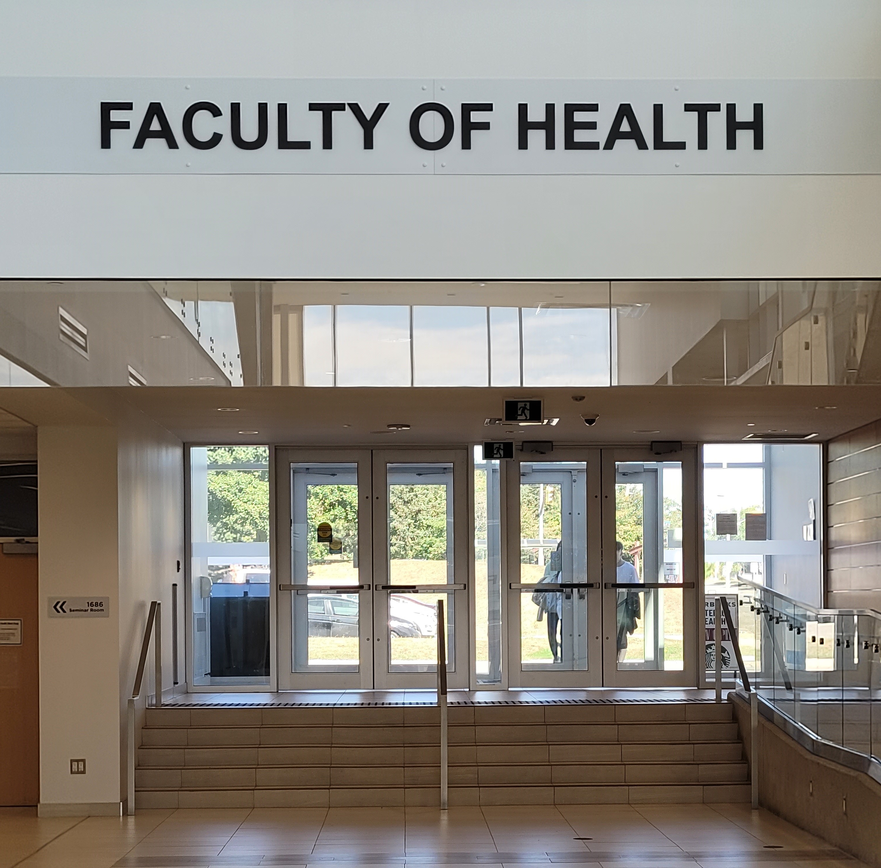 Inside the University of Waterloo. The Faculty of Health sign is above a set of doors with stairs and an accessible ramp.